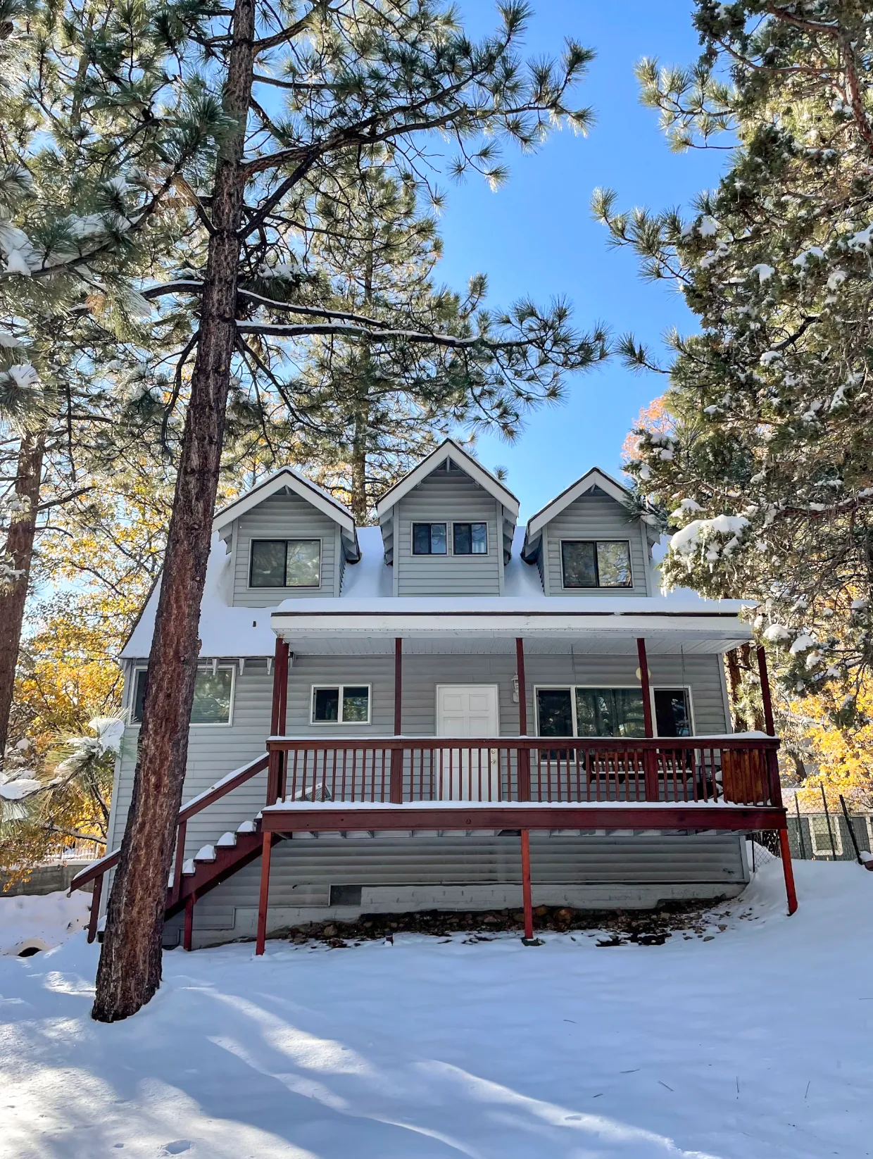 Two-story mountain home with three dormers and a covered porch, surrounded by pines after a snowfall.