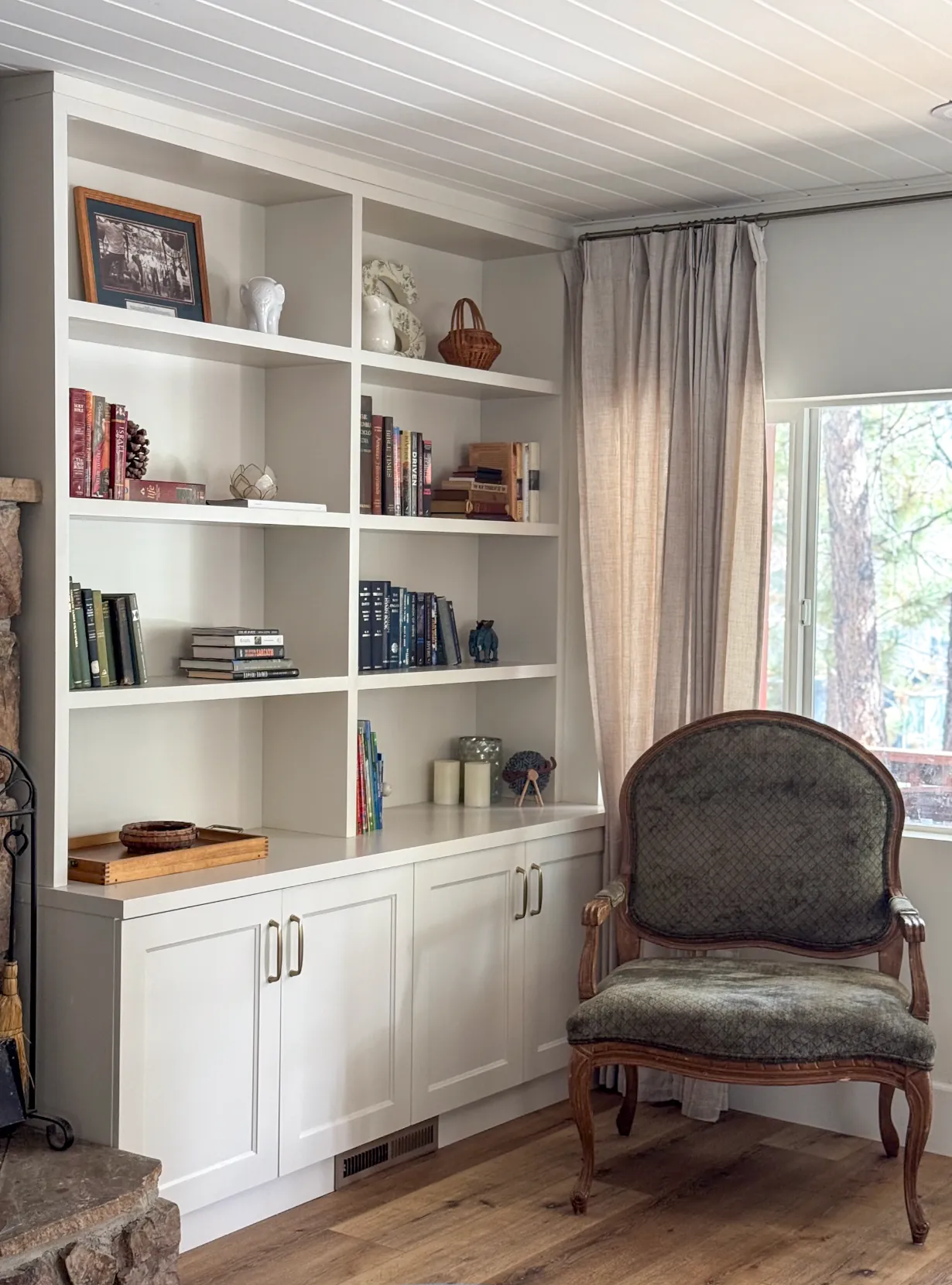 Living room with white built-in shelving, an upholstered antique armchair, and a window looking out to pine trees.